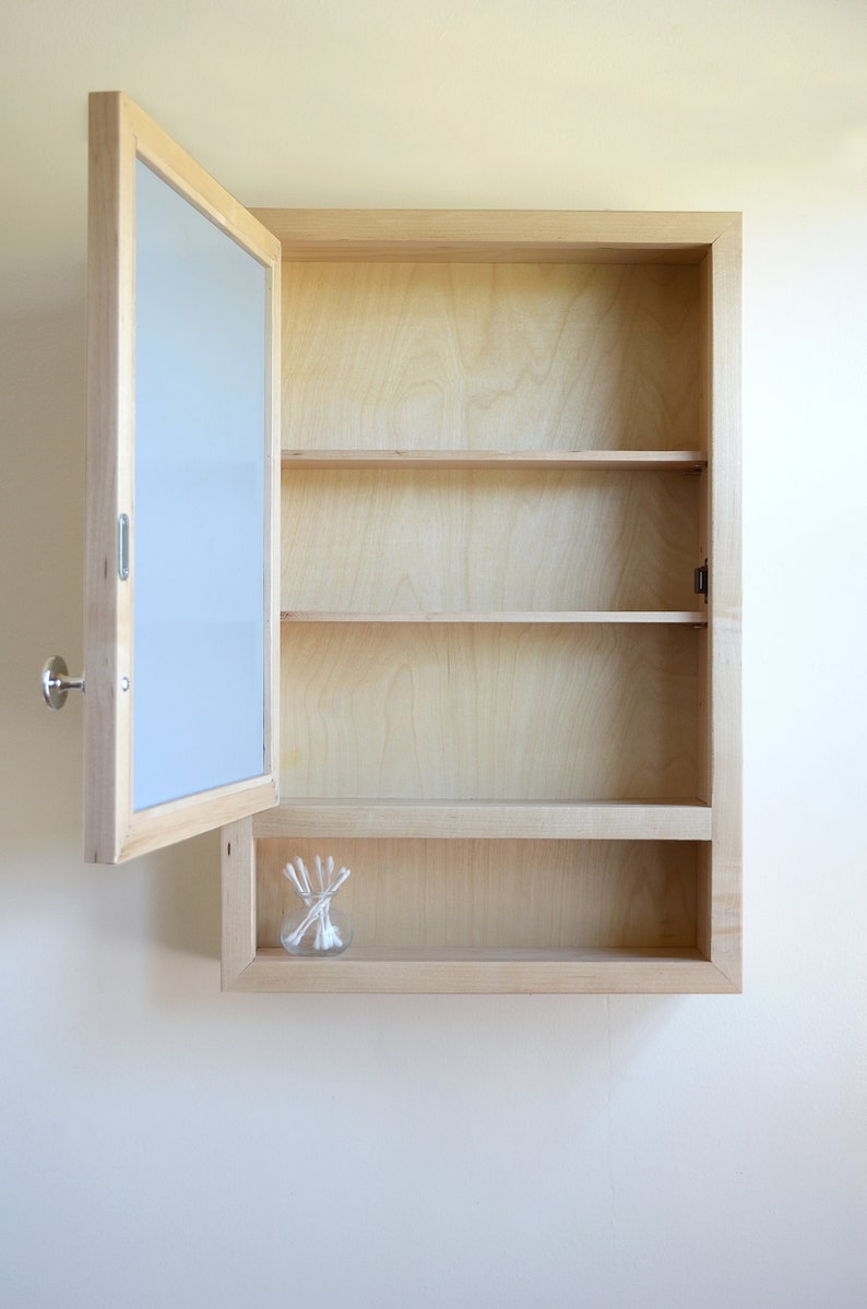 May include: A light-colored wooden medicine cabinet with a mirror door, partially open. The cabinet has three shelves and a small glass jar with cotton swabs. The cabinet is mounted on a white wall.