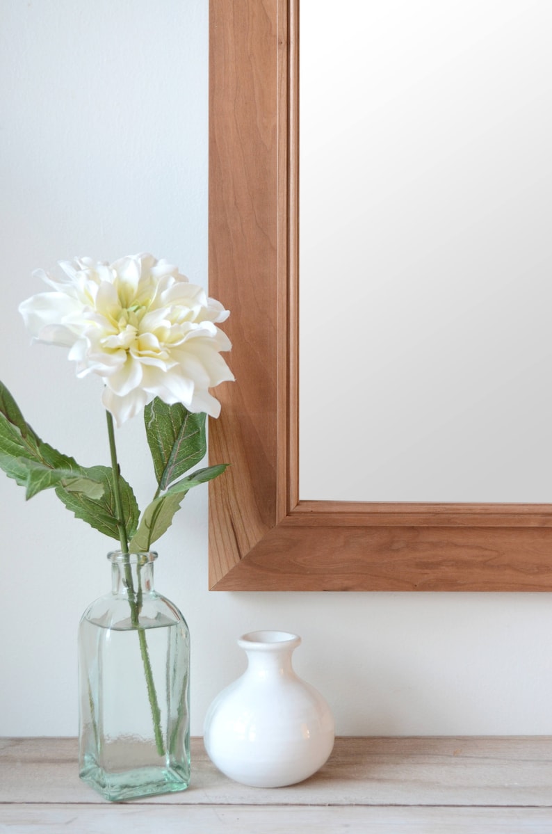 Puede incluir: Una flor blanca en un jarr&oacute;n de vidrio transparente con un jarr&oacute;n de cer&aacute;mica blanca a su lado. El jarr&oacute;n est&aacute; sobre una superficie de madera delante de un espejo con marco de madera.