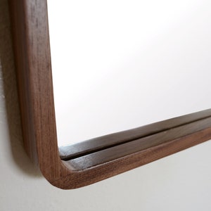 Puede incluir: Primer plano de la esquina inferior derecha de un espejo rectangular con un marco de madera oscura. El marco est&aacute; curvado en el borde exterior.