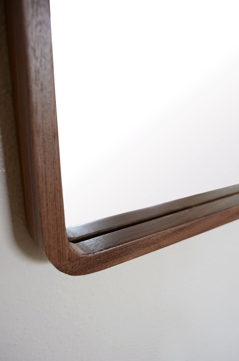 Puede incluir: Primer plano de la esquina inferior derecha de un espejo rectangular con un marco de madera oscura. El marco est&aacute; curvado en el borde exterior.