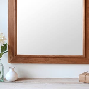 Puede incluir: Un espejo con marco de madera con una flor blanca en un jarr&oacute;n de vidrio transparente y una caja de regalo envuelta en una superficie de madera clara.