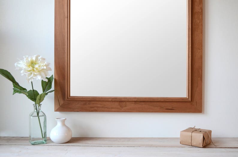 Puede incluir: Un espejo con marco de madera con una flor blanca en un jarr&oacute;n de vidrio transparente y una caja de regalo envuelta en una superficie de madera clara.
