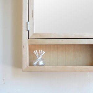May include: A light wood-framed medicine cabinet with a mirror door. The cabinet has a small shelf holding a clear glass jar filled with white cotton swabs. The cabinet is mounted on a white wall.
