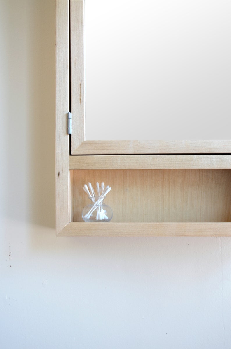 May include: A light wood-framed medicine cabinet with a mirror door. The cabinet has a small shelf holding a clear glass jar filled with white cotton swabs. The cabinet is mounted on a white wall.