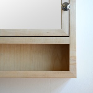 May include: A light-colored wooden medicine cabinet with a mirrored door and a small, round silver knob. The cabinet has a recessed shelf below the door. The wood grain is visible, and the cabinet is mounted on a white wall.