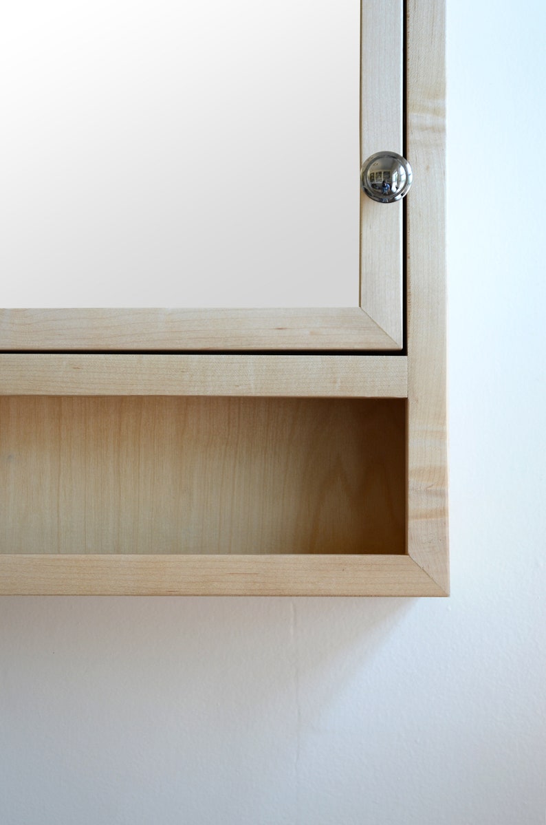 May include: A light-colored wooden medicine cabinet with a mirrored door and a small, round silver knob. The cabinet has a recessed shelf below the door. The wood grain is visible, and the cabinet is mounted on a white wall.