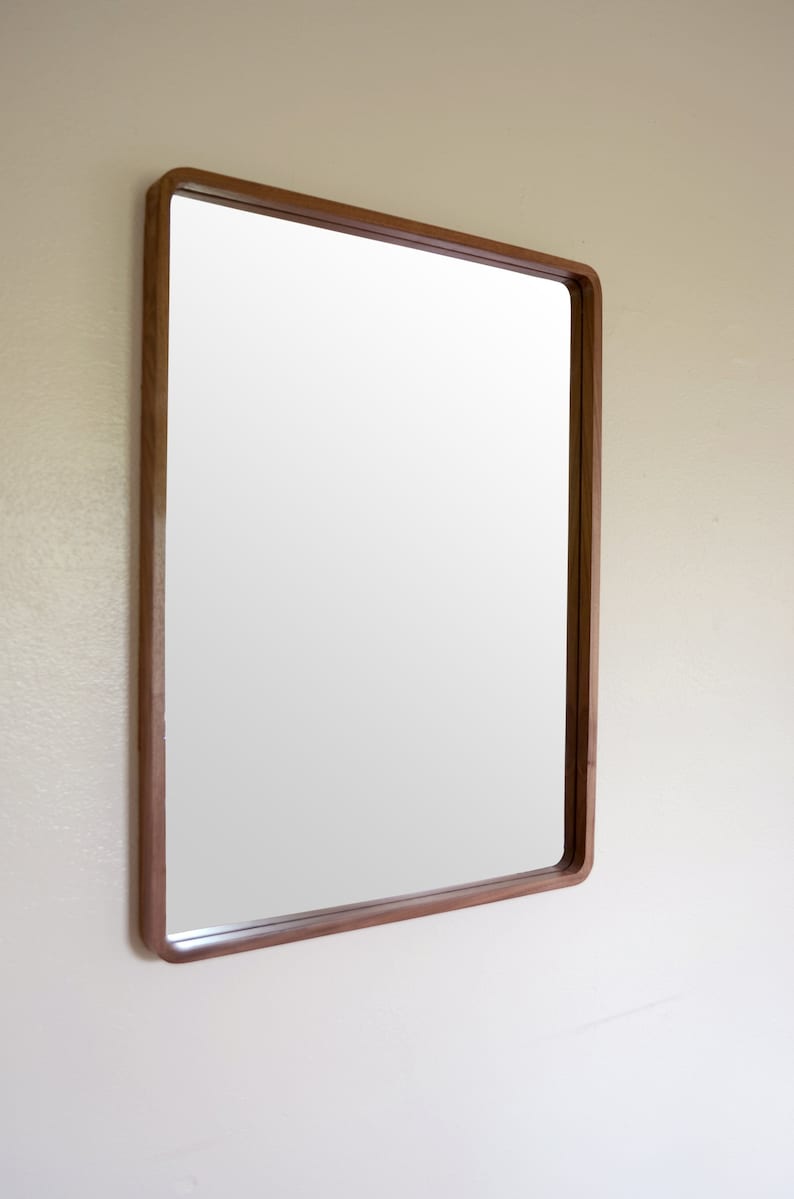 Puede incluir: Un espejo rectangular con un marco de madera marr&oacute;n. El espejo est&aacute; colgado en una pared blanca.