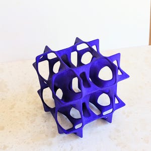 May include: A vibrant purple, geometric sculpture with a cube-like structure. The object features a series of interconnected, open shapes, creating a lattice-like design. The sculpture is made of a solid material and sits on a light-colored surface.