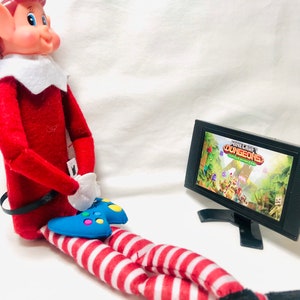 May include: A red and white striped elf doll sitting on a white surface holding a blue video game controller. The elf is looking at a black television screen displaying the video game "Minecraft Dungeons".