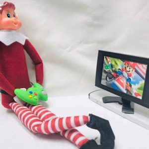 May include: A red and white striped elf doll is sitting on a white surface, holding a green video game controller. The elf is wearing a red and white striped outfit and a red hat. The elf is looking at a small black television screen that is playing a video game. The television screen is on a white stand.