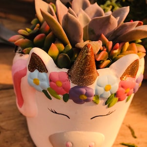 May include: A white unicorn-shaped planter pot filled with a variety of succulents. The pot features a gold glitter horn and ears, a floral crown, and closed eyes. The succulents have green, pink, and purple hues.