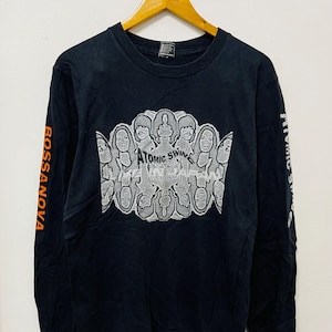 Könnte beinhalten: Schwarzes Langarmshirt mit einem weißen Grafikdruck einer Band namens "Atomic Swing". Die Gesichter der Bandmitglieder wiederholen sich in einem kreisförmigen Muster. Die Ärmel haben die Worte "Bossa Nova" und "Atomic Swing" in Orange gedruckt.