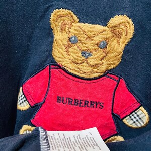 Pu&ograve; includere: Una maglietta blu navy con un orsacchiotto di peluche marrone ricamato che indossa una maglietta rossa con il testo "BURBERRYS" su di essa. La maglietta ha un'etichetta di cura bianca attaccata.