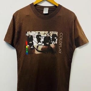 May include: A brown short-sleeve t-shirt featuring a graphic print of the band Coldplay. The rectangular graphic shows four individuals in dark clothing sitting on a floor with cables, accompanied by the text COLDPLAY and a vertical color bar.