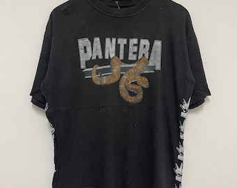 Puede incluir: Camiseta negra con un logotipo blanco de "Pantera" y un gráfico marrón oxidado de una serpiente. La camiseta tiene una impresión blanca de un diseño repetido en la manga derecha.