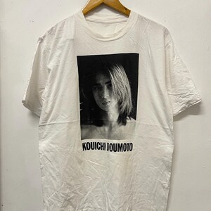 May include: White t-shirt with a black and white photo of a person with long hair. The text "Kouichi Doumoto" is printed below the photo.