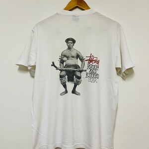 May include: White t-shirt with a graphic of a man holding a weapon, with the text "Stussy Rough and Rugged Gear" in red and black. The shirt is hanging on a wooden hanger.
