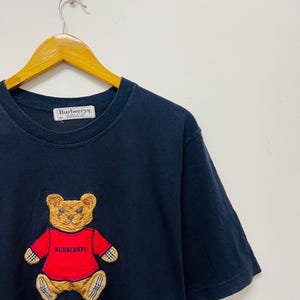 Pu&ograve; includere: Maglietta nera con un orsacchiotto di peluche marrone che indossa una maglietta rossa con il testo "Burberrys" ricamato. La maglietta ha un'etichetta bianca con il testo "Burberrys" su di essa.