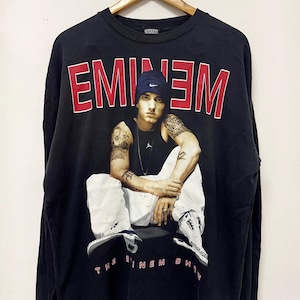 Op de afbeelding: Zwart shirt met lange mouwen met een afbeelding van Eminem in een blauwe beanie en een witte broek. Het woord "EMINEM" is in rode blokletters gedrukt, met "THE EMINEM SHOW" eronder.