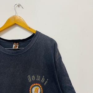 May include: Dark gray t-shirt with a crew neck and short sleeves. The shirt features an embroidered design with the word "Jombi" above a circular emblem with a "J" inside. The emblem is surrounded by a yellow and red sunburst design.
