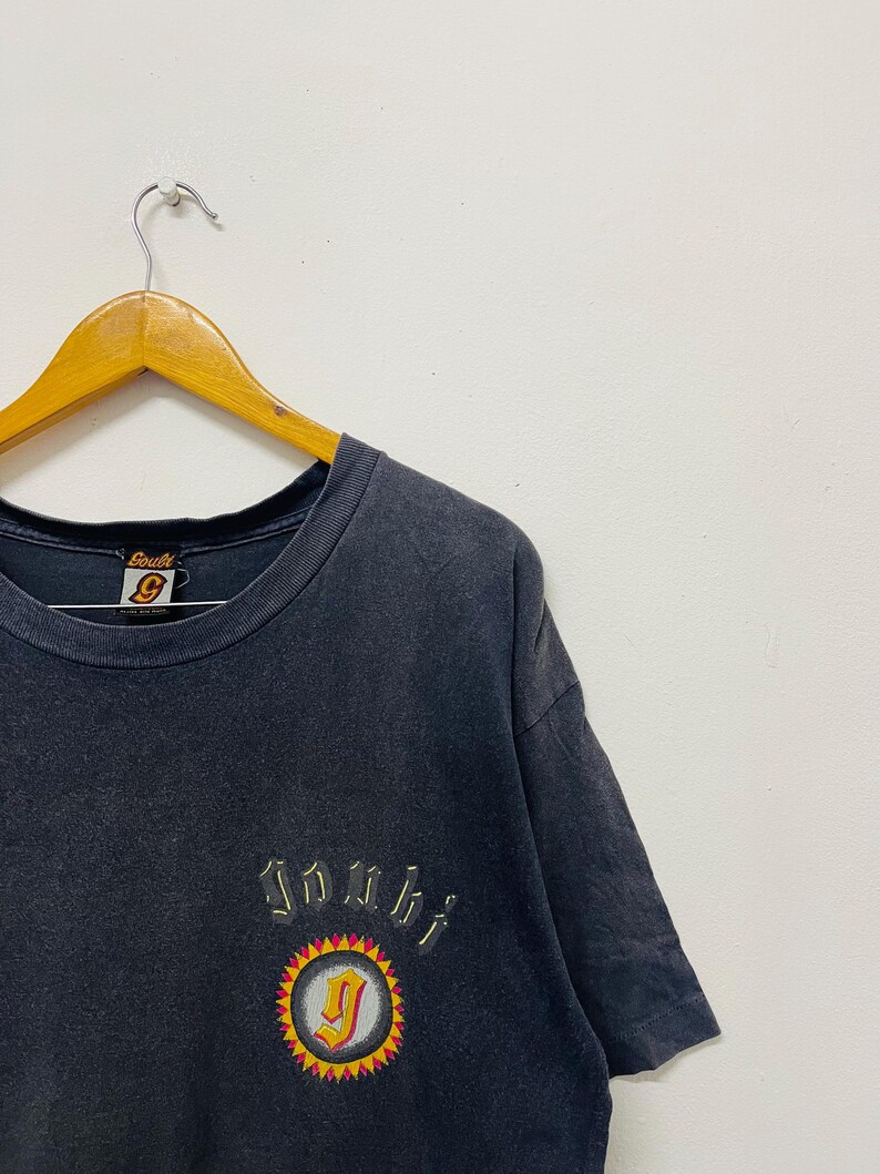 May include: Dark gray t-shirt with a crew neck and short sleeves. The shirt features an embroidered design with the word "Jombi" above a circular emblem with a "J" inside. The emblem is surrounded by a yellow and red sunburst design.