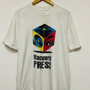 May include: White t-shirt featuring a black and white graphic of a cube with a red, yellow and blue design. The cube has an eye and a speaker on each side. The text "Macworld Press" is printed below the cube.