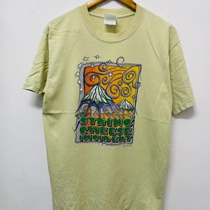 Puede incluir: Una camiseta verde claro con un estampado gráfico de una escena de montaña y el texto "The String Cheese Incident".