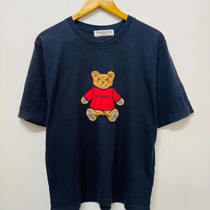 Pu&ograve; includere: Una maglietta nera con un orsacchiotto di peluche ricamato in marrone e rosso che indossa una camicia rossa con la scritta "Burberry's". La maglietta ha un'etichetta bianca con la scritta "Burberry's" su di essa.