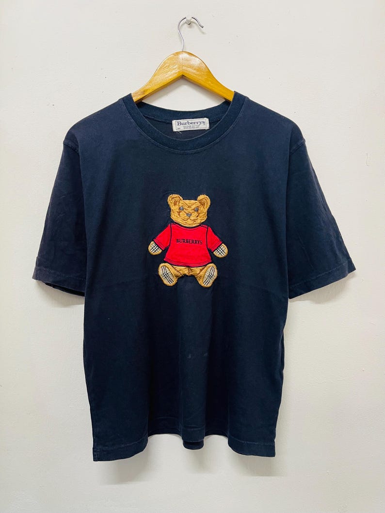 Pu&ograve; includere: Una maglietta nera con un orsacchiotto di peluche ricamato in marrone e rosso che indossa una camicia rossa con la scritta "Burberry's". La maglietta ha un'etichetta bianca con la scritta "Burberry's" su di essa.