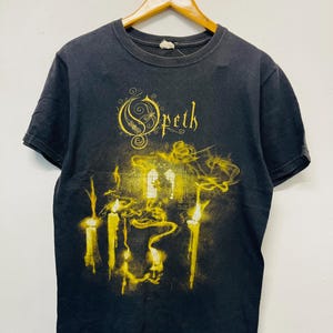 May include: Black t-shirt with a yellow graphic of candles and the band name "Opeth". The design features stylized flames and the band's logo in a gothic font. The shirt is hanging on a wooden hanger.