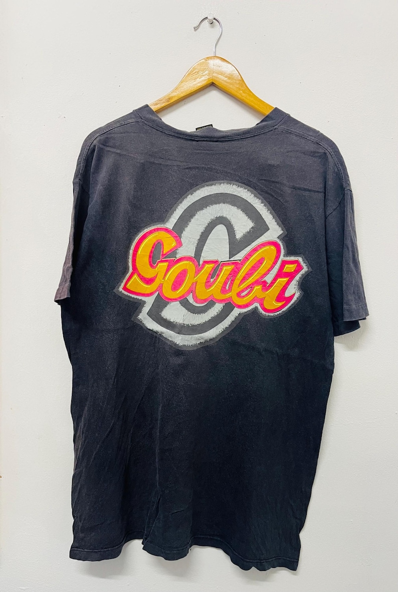 May include: Black t-shirt with a graphic design featuring the word "Goubi" in yellow and pink, outlined in white, set against a gray and white circular design. The shirt is hanging on a wooden hanger.