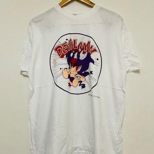May include: White t-shirt with a cartoon image of Devilman in a purple and red costume. The image is surrounded by a blue circle with the text "Devilman" in red and white letters. The text "Comic Plans" is printed in black at the bottom of the image.
