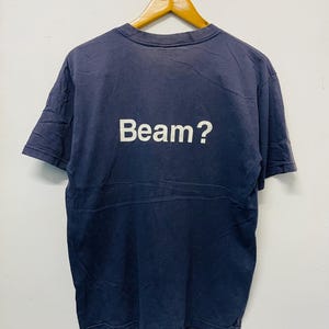 Puede incluir: Una camiseta azul marino con la palabra "Beam?" impresa en blanco. La camiseta está colgada de una percha de madera sobre un fondo blanco. La camiseta parece ser vintage.