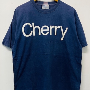 Puede incluir: Camiseta azul marino con la palabra "Cherry" en letras blancas grandes en el pecho. La camiseta está colgada en una percha de madera sobre un fondo blanco. Tiene mangas cortas y cuello redondo.