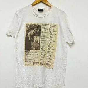 Puede incluir: Camiseta blanca con una imagen en sepia de Morrissey y texto. La imagen es un collage de una fotografía y texto de un periódico. La camiseta está colgada en una percha de madera.
