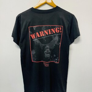 May include: Black t-shirt with a red and black graphic on the back. The graphic includes the word "WARNING!" in red, along with a black and white image and text that reads "The CD-ROM Disc Contains Information No One Wants You to Know About."