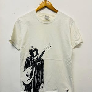 May include: Off-white t-shirt featuring a black and white graphic of a person holding a guitar. The person is wearing a striped suit. The shirt is hanging on a wooden hanger.