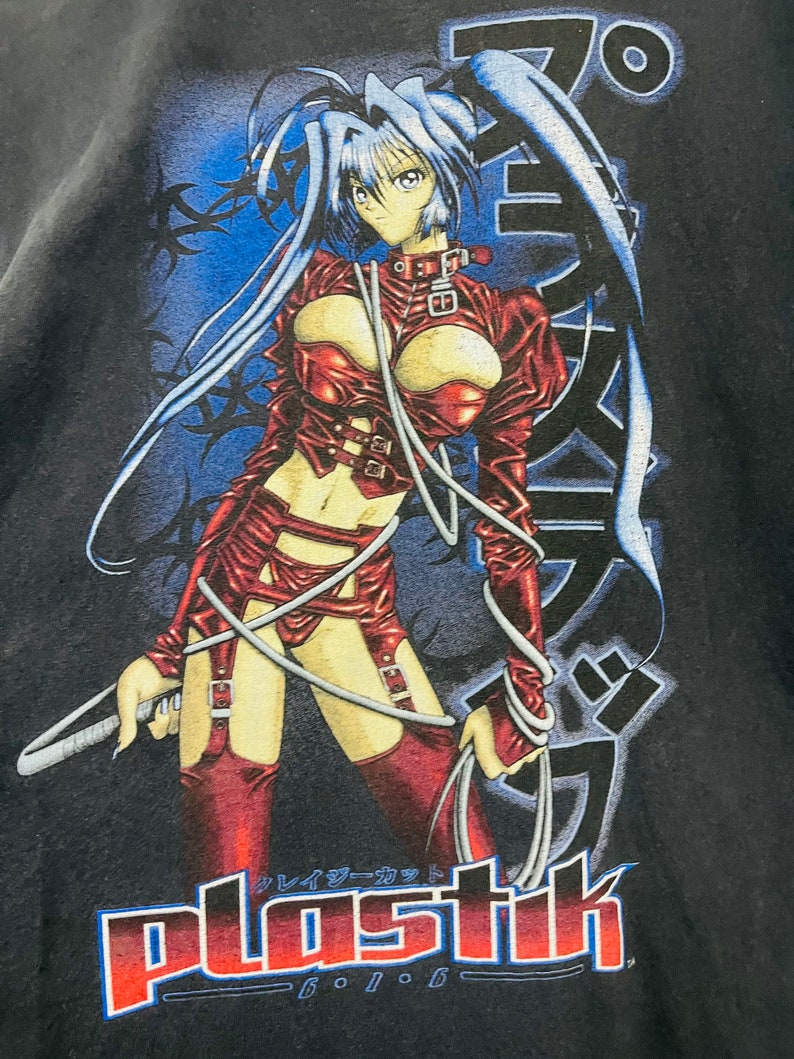 May include: A black t-shirt with a graphic print of an anime character in a red leather outfit. The character is holding a whip and has long blue hair. The text "plastik" is printed in red and blue below the character.