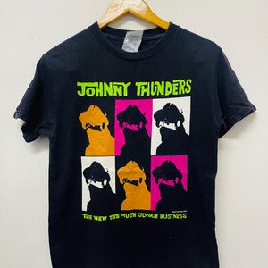 Puede incluir: Camiseta negra con un gráfico verde y blanco de Johnny Thunders. El gráfico presenta una imagen repetida de la cara de Johnny Thunders en una silueta negra con un sombrero blanco y un fondo rosa, naranja o blanco. El texto "Johnny Thunders" está en verde y el texto "The New Too Much Junkie Business" está en verde debajo del gráfico.