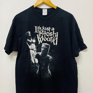 Peut inclure: T-shirt noir avec un graphique blanc d'un chevalier et le texte "It's just a flesh wound."