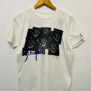 Puede incluir: Camiseta blanca con un diseño gráfico de rectángulos negros con huellas de manos. La palabra "Schimmel" está impresa en azul en cursiva debajo de los rectángulos. La camiseta está colgada en una percha de madera.
