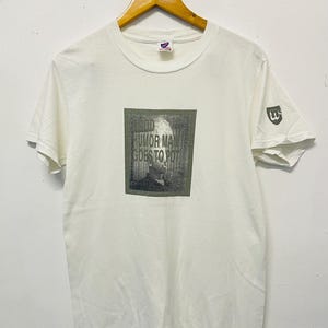 May include: White t-shirt with a printed graphic. The graphic features the text "GOOD HUMOR MAN GOES TO POT" above a grayscale image of a person. The shirt has short sleeves and a small logo on the sleeve.