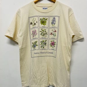 Peut inclure: T-shirt crème avec une grille de neuf illustrations botaniques, chacune représentant une espèce végétale différente. Le texte "Native Plants of Canada" est imprimé en dessous. Le t-shirt est suspendu à un cintre en bois.
