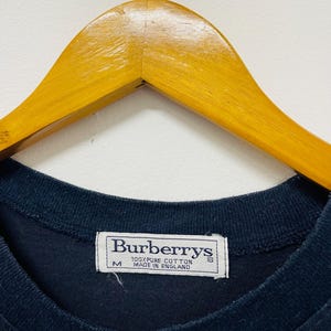 Pu&ograve; includere: Una t-shirt nera Burberrys con un'etichetta bianca che recita "100% puro cotone Prodotto in Inghilterra" e un'etichetta di taglia "M".