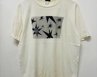 Può includere: Una maglietta bianca con una stampa grafica in bianco e nero di stelle. La grafica è centrata sulla maglietta e ha un cerchio blu al centro.