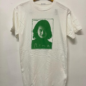 Puede incluir: Camiseta blanca con un estampado gráfico verde de la cara de una mujer con el texto "KYONJUB" y "Rina" en ella.