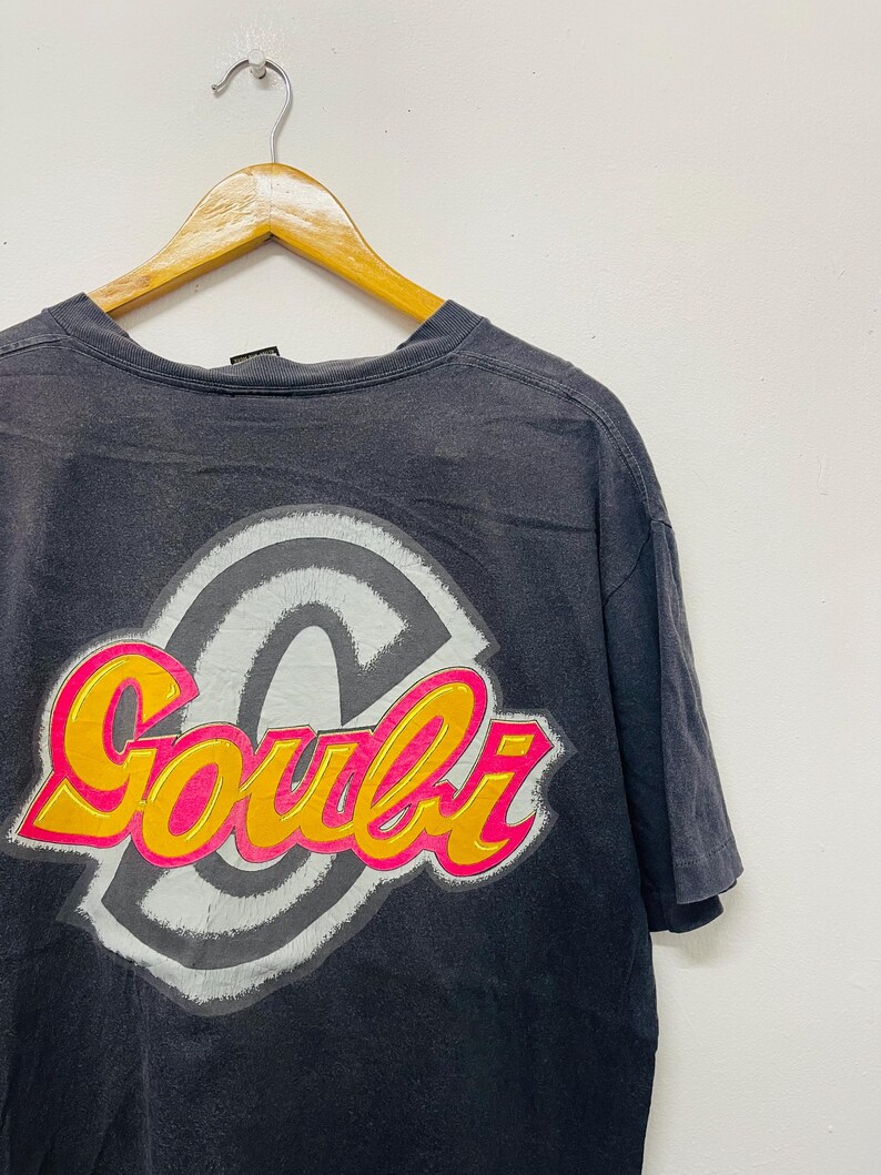 May include: Dark gray t-shirt with a large graphic on the back. The graphic features the word "Goubi" in a vibrant pink and yellow font, outlined in orange, set against a gray and white circular design.