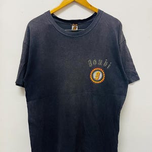May include: A vintage black t-shirt with a small embroidered design. The design features the word "Joshi" above a circular emblem with a yellow sun and a letter "J". The shirt is hanging on a wooden hanger.