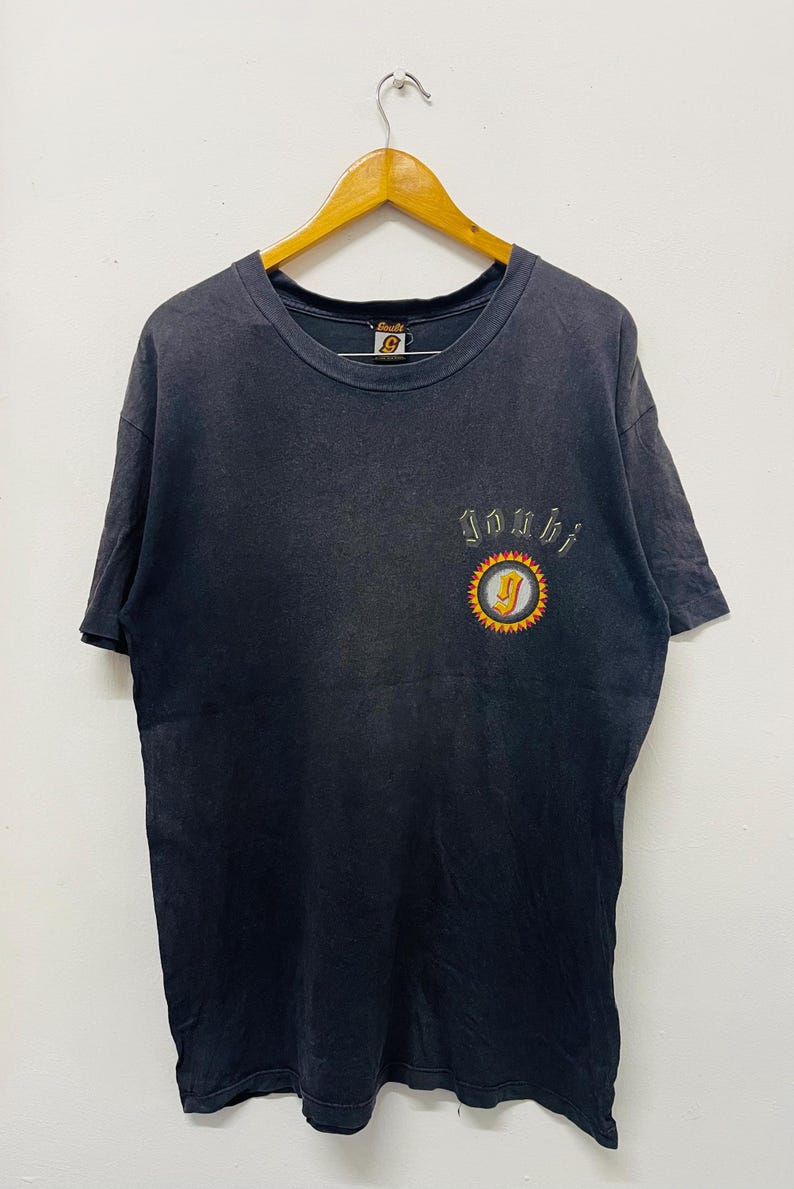 May include: A vintage black t-shirt with a small embroidered design. The design features the word "Joshi" above a circular emblem with a yellow sun and a letter "J". The shirt is hanging on a wooden hanger.