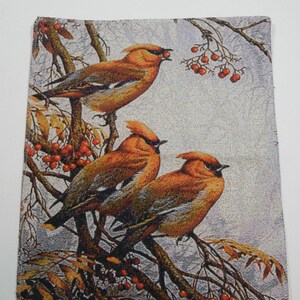 May include: A tapestry wall hanging featuring three orange and brown birds perched on a branch with red berries. The birds have black eyes and beaks. The background is a light gray with a subtle pattern.